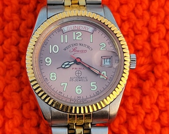 Puede incluir: Un reloj de pulsera de dos tonos, dorado y plateado, con una esfera rosa. La esfera del reloj tiene marcadores de hora en números romanos negros y una pantalla del día de la semana. La correa del reloj es una correa de eslabones de metal de dos tonos, dorado y plateado. La esfera del reloj también tiene el texto "West End Watch Co. Sowan" y "Prima Automatic 25 Jewels".