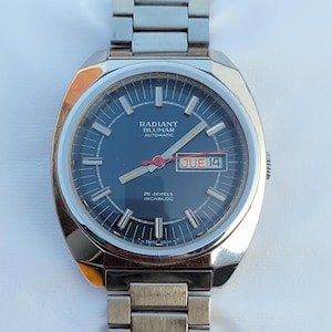 May include: A silver Radiant Blumar automatic wristwatch with a black face, silver hands, and a silver metal band. The watch face has a date window that reads "JUE 14". The watch face also has the text "25 JEWELS INCABLOC" and "SWISS MADE T".