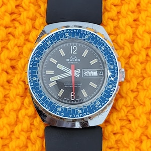 May include: A black wristwatch with a blue and orange bezel. The watch face is black with white markings and a red second hand. The watch face also has a date window and a day of the week window. The watch is labelled "BULER ESPECIAL" and "SHOCKRISTANT ANTIMAGNETIC".