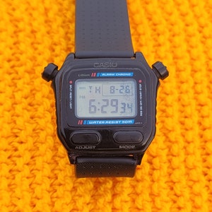 May include: A black Casio digital watch with a black band. The watch face displays the time, date, and day of the week. The watch is water resistant to 50 meters. The watch is on a yellow knitted surface.