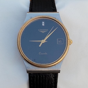 May include: A silver and gold Longines watch with a black face and a black leather strap. The watch has a diamond at the 12 o'clock position and the words "Longines" and "Quartz" printed on the face.
