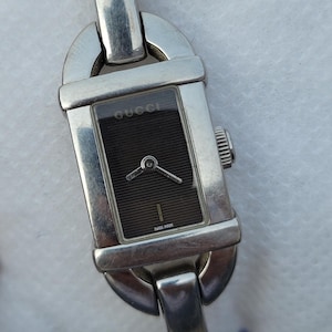 May include: A rectangular Gucci watch with a silver strap and a black face. The watch face has the Gucci logo and the words "Swiss Made". The watch has a silver crown and a silver clasp.