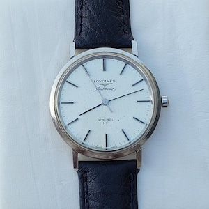 May include: A silver Longines Admiral HF automatic wristwatch with a white face and black leather band. The watch face features black hour markers and hands, with the Longines logo and "Automatic" text. The watch is set against a white background.
