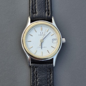 May include: An Omega Seamaster wristwatch featuring a silver-toned dial with vertical pinstripes, gold-toned hour markers, and a date window at the three o'clock position. The watch has a silver-toned case with a gold-toned bezel and a black leather strap.
