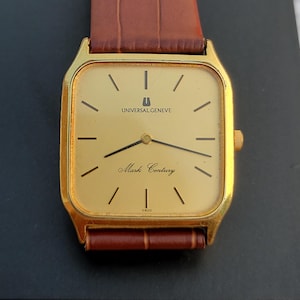 May include: A gold-plated Universal Geneve Mark Century wristwatch with a rectangular face and brown leather strap. The watch face is gold with black hour markers and hands. The strap is dark brown, textured leather.