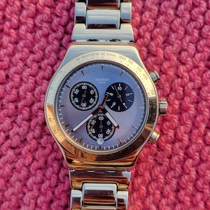 May include: A gold-toned Swatch chronograph watch with a light gray face and black sub-dials. The watch features a tachymeter bezel and a stainless steel band. The watch is displayed on a textured pink surface.