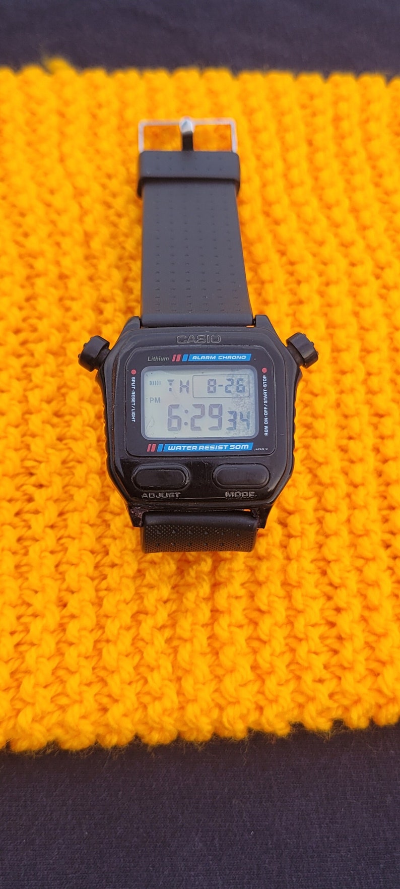Puede incluir: Un reloj digital Casio negro con una correa negra. La esfera del reloj muestra la hora, la fecha y el d&iacute;a de la semana. El reloj es resistente al agua hasta 50 metros. El reloj est&aacute; sobre una superficie de punto amarillo.