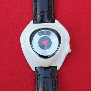 Puede incluir: Un reloj de pulsera blanco vintage con una correa de cuero negro. La esfera del reloj presenta un dise&ntilde;o circular con un tri&aacute;ngulo rojo y los n&uacute;meros 7, 8 y 9. El reloj tiene una est&eacute;tica retro &uacute;nica.
