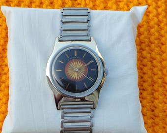 Puede incluir: Un reloj de pulsera plateado con una esfera negra y un diseño de sol dorado. El reloj tiene una correa de metal plateado.
