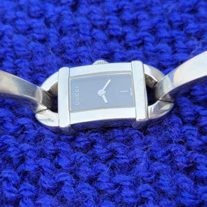 May include: A silver wristwatch with a rectangular black face and silver hands. The watch has a curved band that extends on either side of the watch face. The watch is displayed on a textured blue surface.