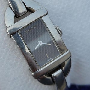 May include: A silver Gucci wristwatch with a rectangular dark grey face. The watch has a silver band and the Gucci logo is visible on the watch face. The words "Swiss Made" are printed on the watch face.