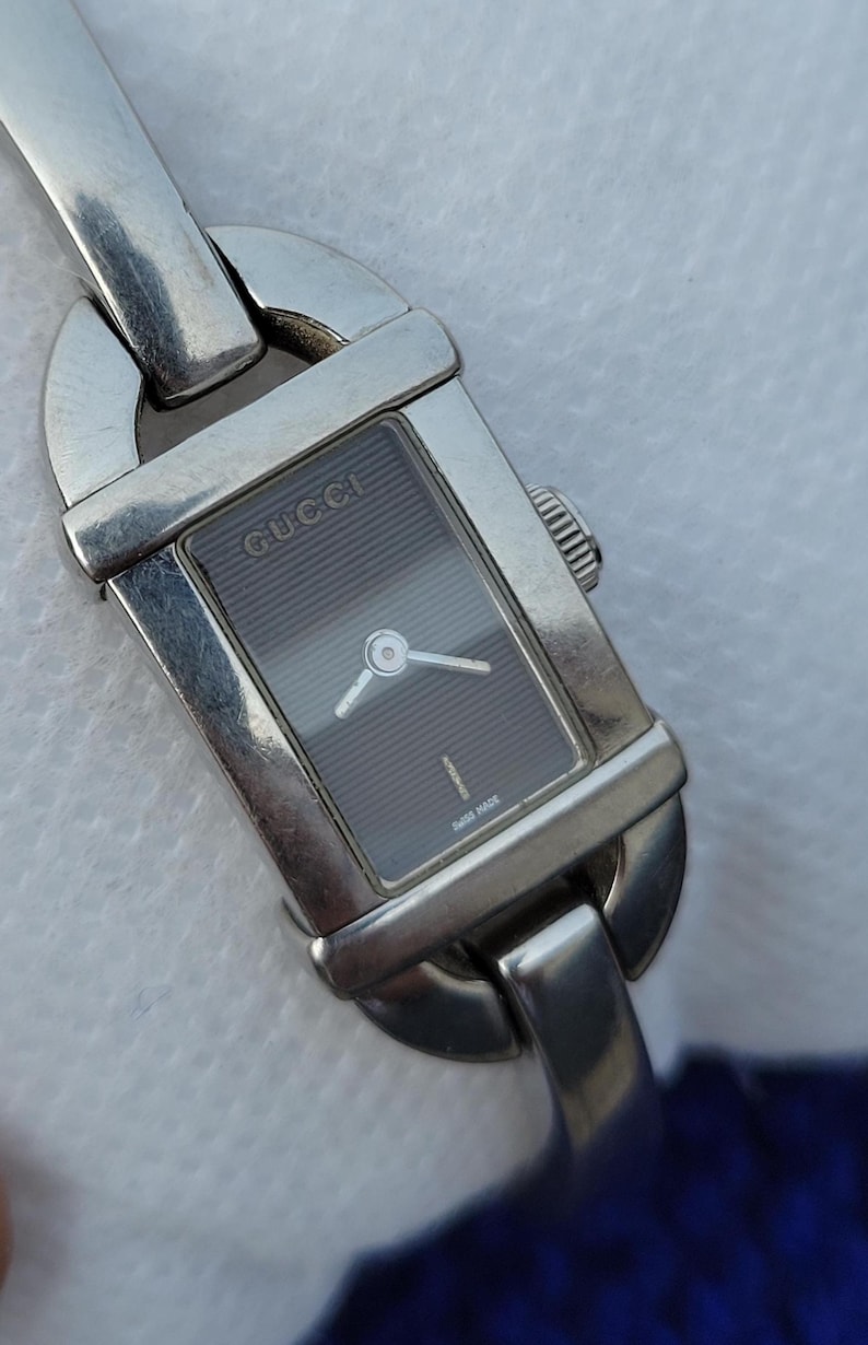 May include: A silver Gucci wristwatch with a rectangular dark grey face. The watch has a silver band and the Gucci logo is visible on the watch face. The words "Swiss Made" are printed on the watch face.