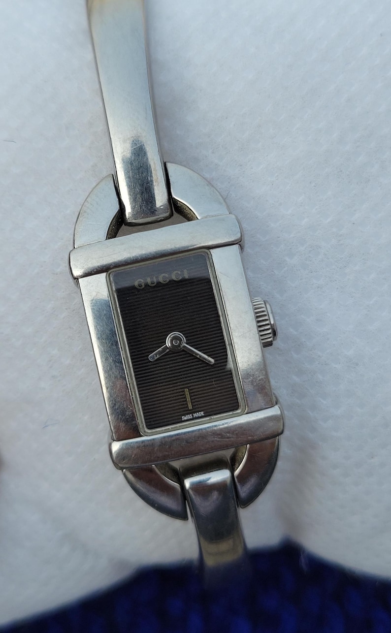 May include: A rectangular Gucci watch with a silver strap and a black face. The watch face has the Gucci logo and the words "Swiss Made". The watch has a silver crown and a silver clasp.