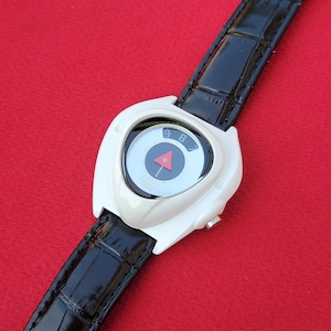 Puede incluir: Un reloj de pulsera blanco con una correa de cuero negro. La esfera del reloj es circular con un tri&aacute;ngulo rojo y una flecha negra. El reloj tiene los n&uacute;meros 8 y 9 en la esfera. La correa es negra y tiene un patr&oacute;n texturizado.