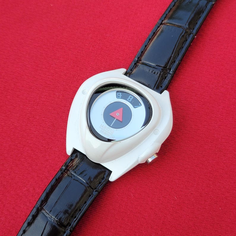 Puede incluir: Un reloj de pulsera blanco con una correa de cuero negro. La esfera del reloj es circular con un tri&aacute;ngulo rojo y una flecha negra. El reloj tiene los n&uacute;meros 8 y 9 en la esfera. La correa es negra y tiene un patr&oacute;n texturizado.