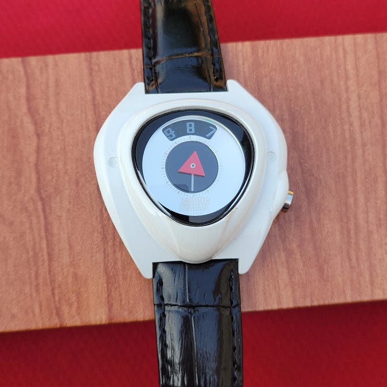 Puede incluir: Reloj de pulsera blanco con correa de cuero negro. La esfera del reloj tiene un dise&ntilde;o &uacute;nico con un elemento circular central, un tri&aacute;ngulo rojo y los n&uacute;meros 8 y 9. El reloj tiene una est&eacute;tica elegante y moderna.