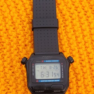 Puede incluir: Un reloj digital Casio negro con una correa negra. La esfera del reloj muestra la hora, la fecha y el d&iacute;a de la semana. El reloj es resistente al agua hasta 50 metros. El reloj tiene un bot&oacute;n negro etiquetado como "ADJUST" y un bot&oacute;n negro etiquetado como "MODE".