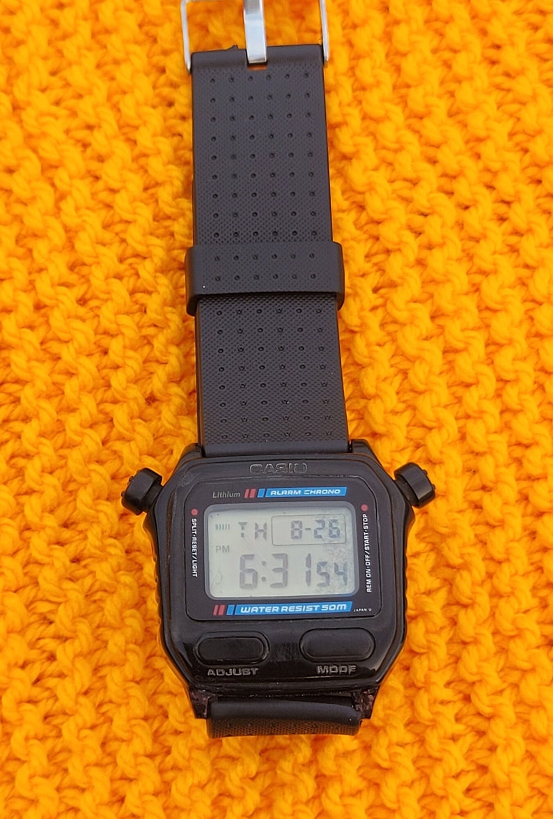Puede incluir: Un reloj digital Casio negro con una correa negra. La esfera del reloj muestra la hora, la fecha y el d&iacute;a de la semana. El reloj es resistente al agua hasta 50 metros. El reloj tiene un bot&oacute;n negro etiquetado como "ADJUST" y un bot&oacute;n negro etiquetado como "MODE".