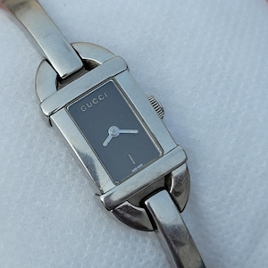 May include: A silver Gucci wristwatch with a rectangular black face. The watch has a silver band and a small dial with hands. The word "GUCCI" is printed on the watch face. The watch is a classic design.