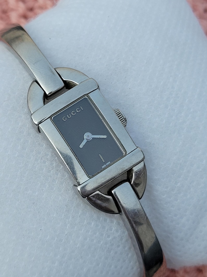 May include: A silver Gucci wristwatch with a rectangular black face. The watch has a silver band and a small dial with hands. The word "GUCCI" is printed on the watch face. The watch is a classic design.
