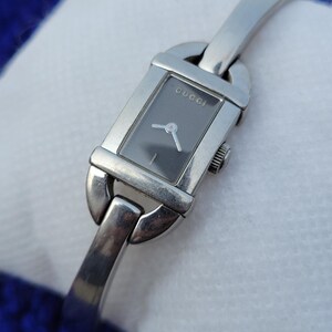May include: A silver Gucci wristwatch with a rectangular dark grey face. The watch has a silver band and a small crown. The word "GUCCI" is printed on the watch face.
