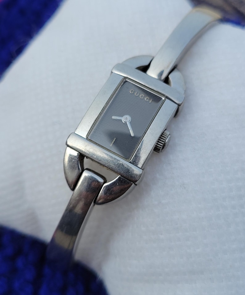 May include: A silver Gucci wristwatch with a rectangular dark grey face. The watch has a silver band and a small crown. The word "GUCCI" is printed on the watch face.