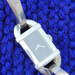 May include: A silver Gucci watch with a rectangular grey face and silver hands. The watch band is also silver and has a unique design. The word "GUCCI" is printed on the watch face. The watch is displayed on a blue textured surface.