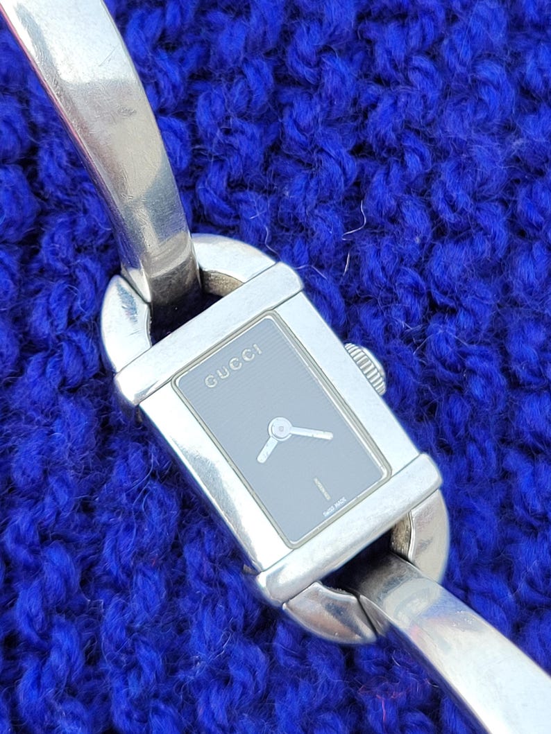 May include: A silver Gucci watch with a rectangular grey face and silver hands. The watch band is also silver and has a unique design. The word "GUCCI" is printed on the watch face. The watch is displayed on a blue textured surface.