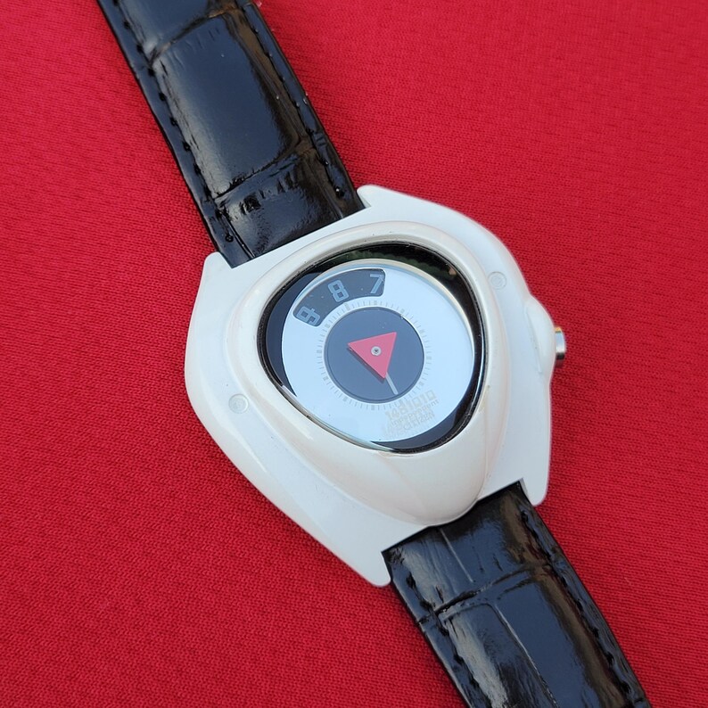 Puede incluir: Un reloj de pulsera blanco con una correa de cuero negro. La esfera del reloj tiene un dise&ntilde;o circular con un tri&aacute;ngulo rojo y los n&uacute;meros 7 y 8. El reloj est&aacute; sobre un fondo rojo.