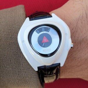 Puede incluir: Reloj de pulsera blanco con un dise&ntilde;o de caja triangular &uacute;nico. La esfera es negra y plateada con un tri&aacute;ngulo rojo y los n&uacute;meros 7, 8 y 9. La correa es de cuero negro.