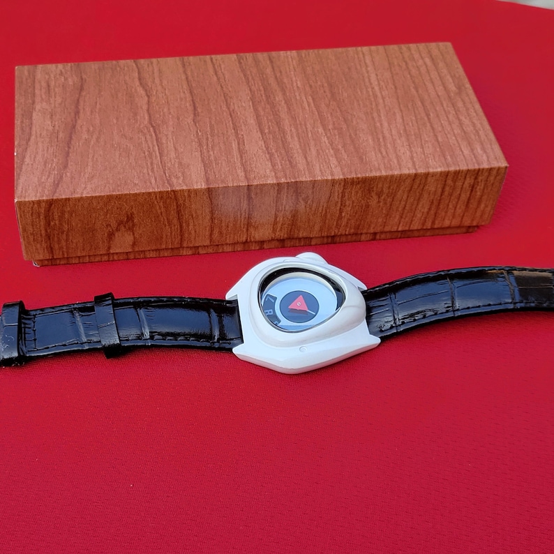 Puede incluir: Un reloj de pulsera blanco con una correa de cuero negro y un dise&ntilde;o de esfera &uacute;nico. La esfera del reloj presenta un c&iacute;rculo azul con un tri&aacute;ngulo rojo. Una caja de madera est&aacute; en el fondo.