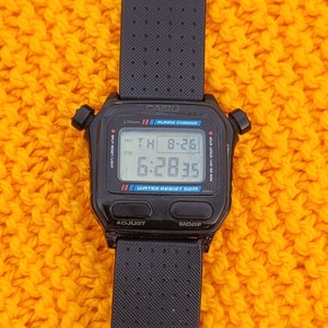 Puede incluir: Un reloj digital Casio negro con una correa negra. La esfera del reloj muestra la hora, la fecha y la alarma. El reloj es resistente al agua hasta 50 metros.