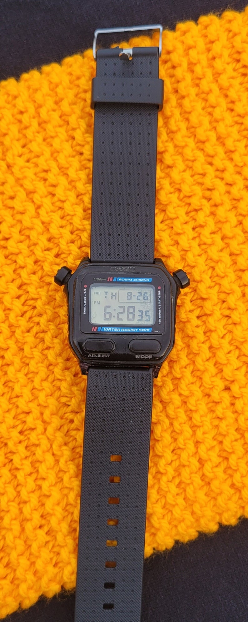 Puede incluir: Un reloj digital Casio negro con una correa negra. La esfera del reloj muestra la hora, la fecha y la alarma. El reloj es resistente al agua hasta 50 metros.