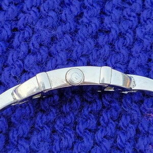 May include: Close-up of a silver wristwatch with a round face and a silver band. The watch face has a small, circular design. The watch is set against a textured, blue background.