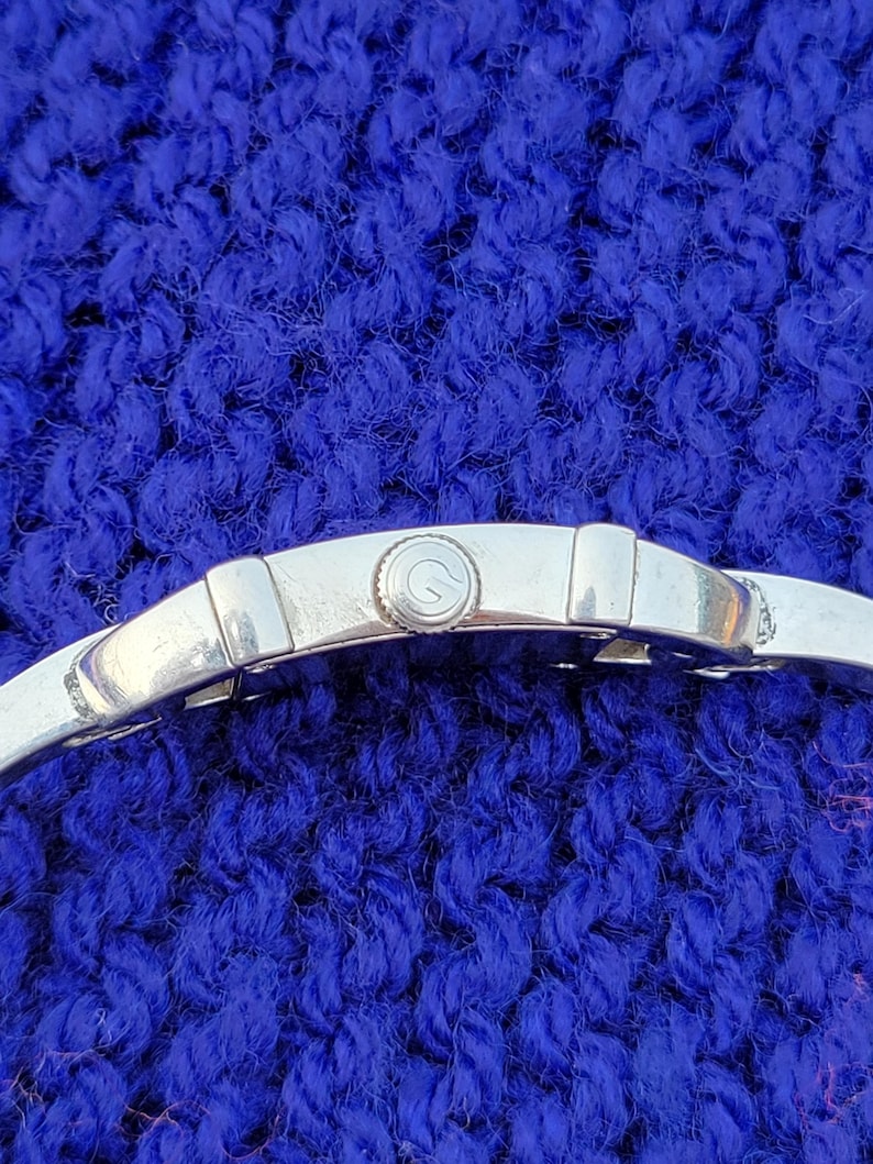 May include: Close-up of a silver wristwatch with a round face and a silver band. The watch face has a small, circular design. The watch is set against a textured, blue background.