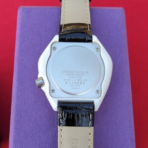 Puede incluir: Parte trasera de un reloj Citizen con caja de pl&aacute;stico blanco y correa de cuero negro. La esfera plateada muestra el texto "CITIZEN WATCH CO. WATER RESIST PLASTICS" y otros detalles. La correa es negra con la parte inferior beige.
