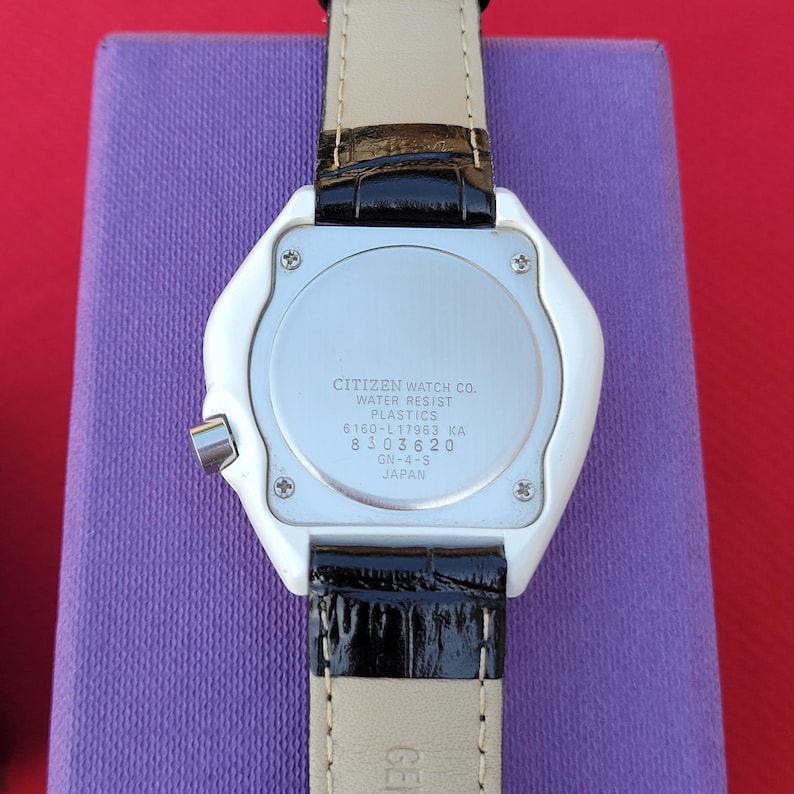 Puede incluir: Parte trasera de un reloj Citizen con caja de pl&aacute;stico blanco y correa de cuero negro. La esfera plateada muestra el texto "CITIZEN WATCH CO. WATER RESIST PLASTICS" y otros detalles. La correa es negra con la parte inferior beige.