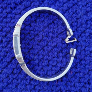 May include: A silver hinged bangle bracelet. The bracelet is open, showing the clasp mechanism. The background is a textured, deep blue fabric.