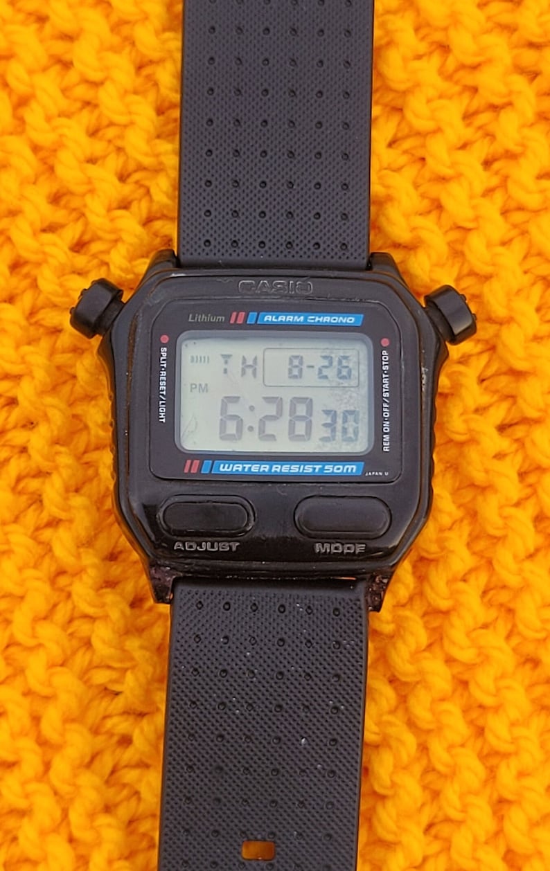 Puede incluir: Un reloj digital Casio negro con una correa negra. La esfera del reloj muestra la hora, la fecha y el d&iacute;a de la semana. El reloj es resistente al agua hasta 50 metros.
