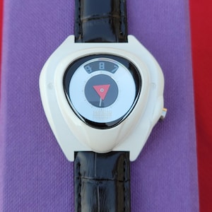 Puede incluir: Un reloj de pulsera blanco con una correa de cuero negro. La esfera del reloj tiene un dise&ntilde;o en blanco y negro con un tri&aacute;ngulo rojo y los n&uacute;meros 8 y 9. El reloj est&aacute; sobre una superficie morada.