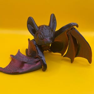 May include: A 3D printed bat figurine with a dark purple color. The bat has its wings spread out and is sitting on a yellow surface.