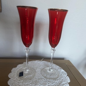 Puede incluir: Dos copas de champán rojas con bordes dorados. Las copas tienen un tallo transparente y adornado y una base transparente. Están sobre un tapete de encaje blanco. Las copas son de color rojo vibrante.