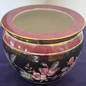 Könnte beinhalten: Ein runder, dekorativer Keramik-Pflanztopf mit einer weiten Öffnung. Der Pflanztopf hat eine schwarze Basis, die mit floralen Mustern in Rosa, Blau und Weiß verziert ist. Der Rand und ein Band in der Mitte sind in einem tiefen Rosaton gehalten, mit goldenen Akzenten.