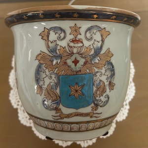 Puede incluir: Jardinera de cerámica blanca con un diseño de escudo detallado. El escudo presenta un escudo azul con una estrella dorada, rodeado de adornos dorados y azules. El borde tiene una banda azul oscuro con estrellas doradas.