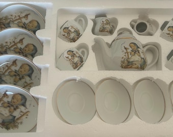 Op de afbeelding: Een wit porseleinen theeservies met gouden rand, bestaande uit een theepot, kopjes, schotels en een melkkan. Elk stuk is versierd met een gedetailleerde illustratie van een kind. De set is gerangschikt in een beschermende witte schuimrubberen inzet.