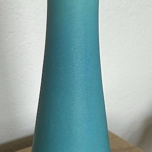 May include: A tall, slender vase with a matte turquoise finish. The vessel features a flared rim that tapers down into a narrow neck, gradually widening toward a circular base. The surface has a consistent, smooth texture in a solid blue-green color.