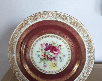 May include: A white porcelain plate with a burgundy rim and gold trim. The center of the plate features a floral design with pink, yellow, and purple flowers.