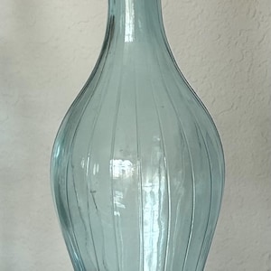 Puede incluir: Una botella alta de vidrio azul claro con tapón. La botella tiene un diseño acanalado y una base redondeada. El tapón también es azul claro y tiene una forma decorativa. La botella mide unos 30 cm de altura.