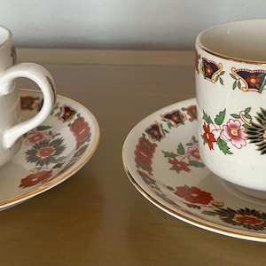May include: Two white teacups and saucers with floral and geometric designs. The cups and saucers have a gold trim. The floral design includes red, yellow, and green flowers. The saucers have a matching floral pattern.