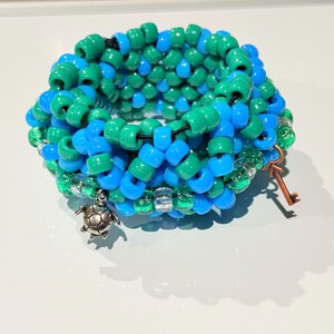 May include: A multi-colored beaded bracelet with a green and blue color scheme. The bracelet has a silver turtle charm and a copper key charm.